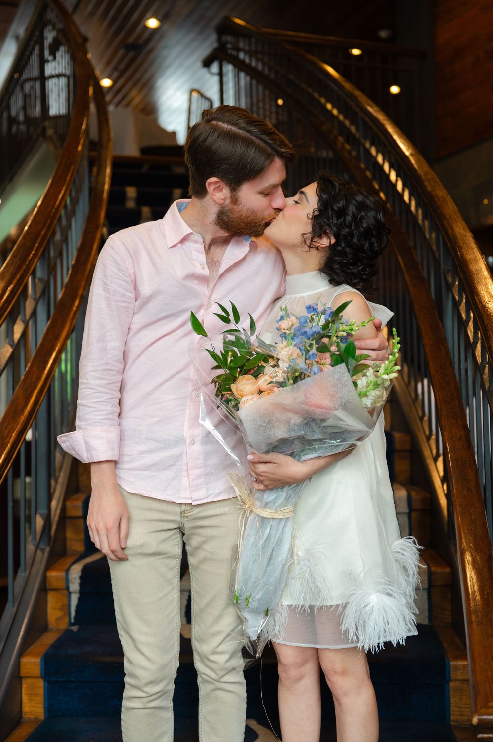 Couple sharing a kiss on elegant staircase, woman holding bouquet of blue and peach flowers, wearing white feathered dress.