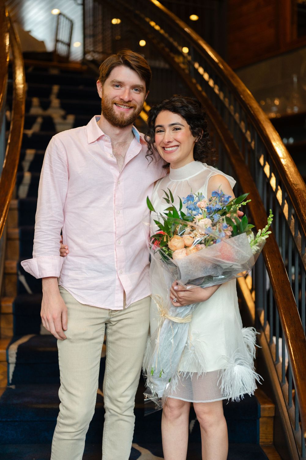 Smiling couple posing on ornate golden staircase, woman in white feathered dress holding blue and peach floral bouquet.