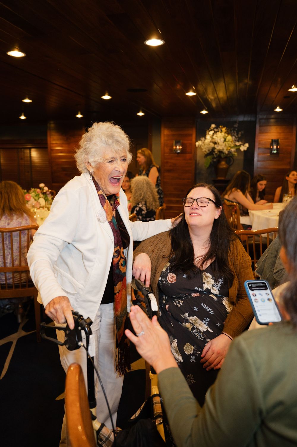 Elderly woman with walker laughs with younger woman in wheelchair at elegant indoor event.