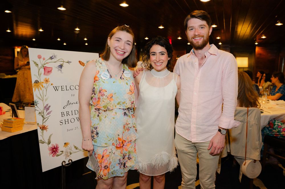Three guests smiling together at a bridal show event, standing near a welcome sign at an indoor venue.