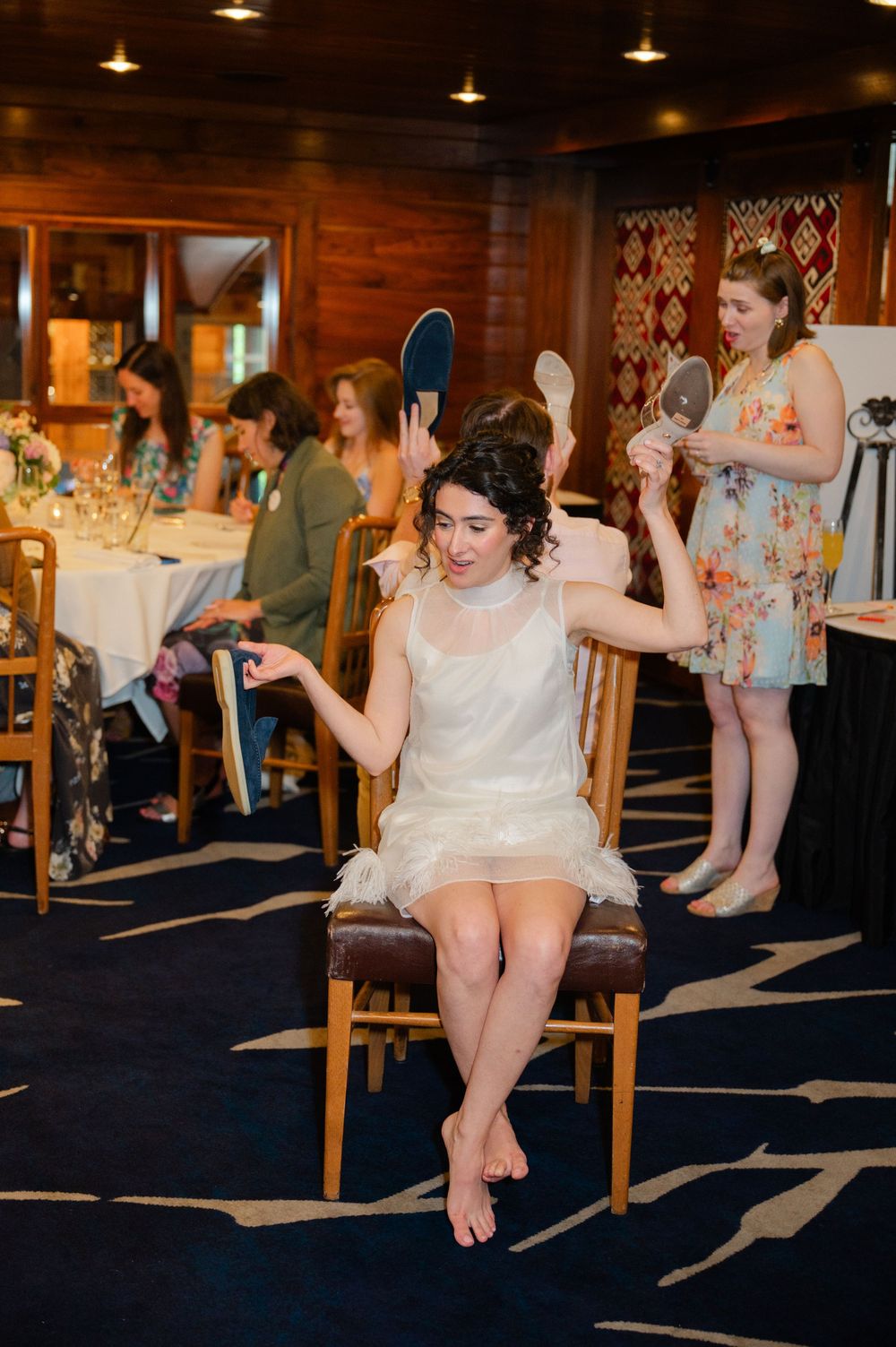 Bride in white dress sits barefoot holding mirror while bridesmaids help with hair and makeup in elegant wood-paneled room.