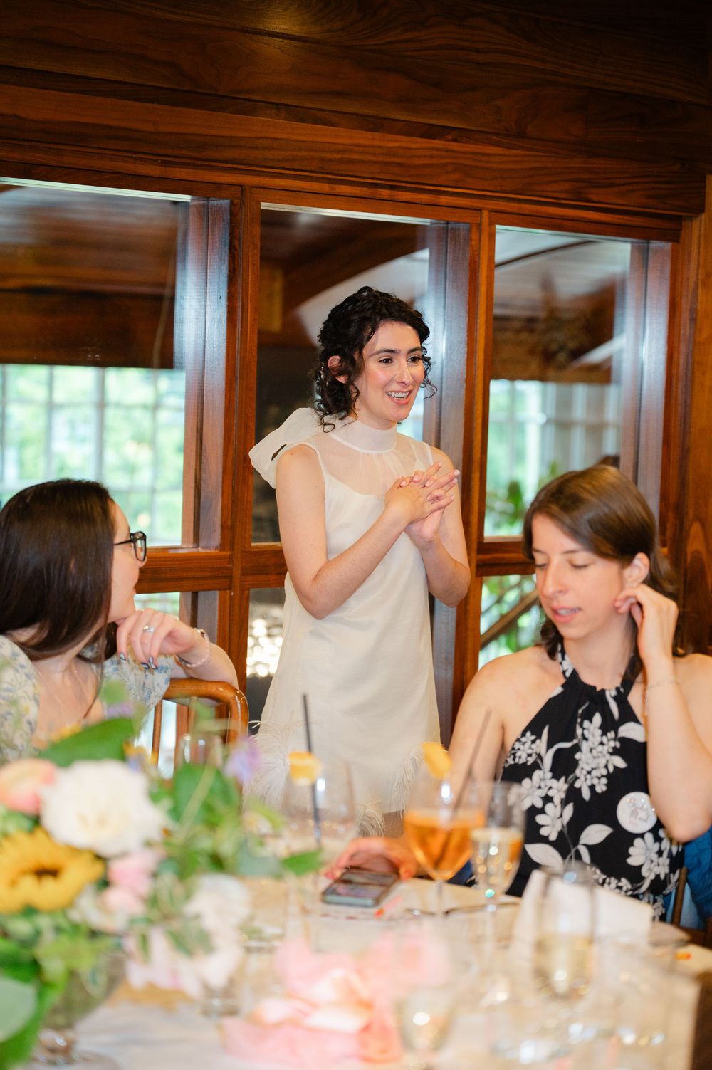 A bride in a white dress smiles and clasps her hands while speaking to guests at an elegant wedding reception dinner.