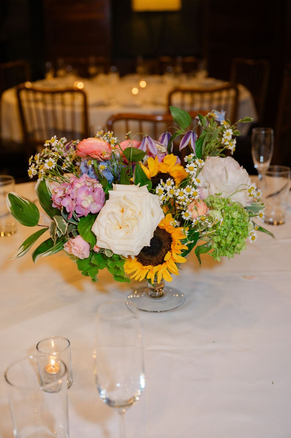 Elegant wedding centerpiece with sunflowers, white roses, pink peonies, and greenery in a glass compote vase on a white tablecloth.
