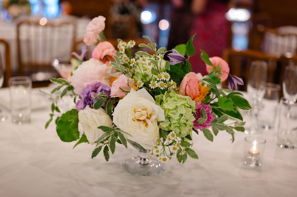 Elegant wedding centerpiece with pink peonies, white roses, purple blooms, and lush greenery in a silver vase on a white tablecloth.