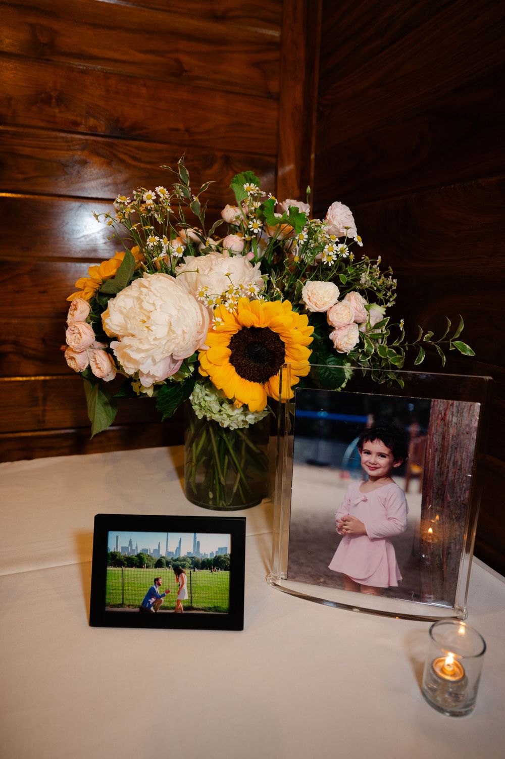 Floral centerpiece with sunflower and white peonies beside framed family photos and candle on white tablecloth.