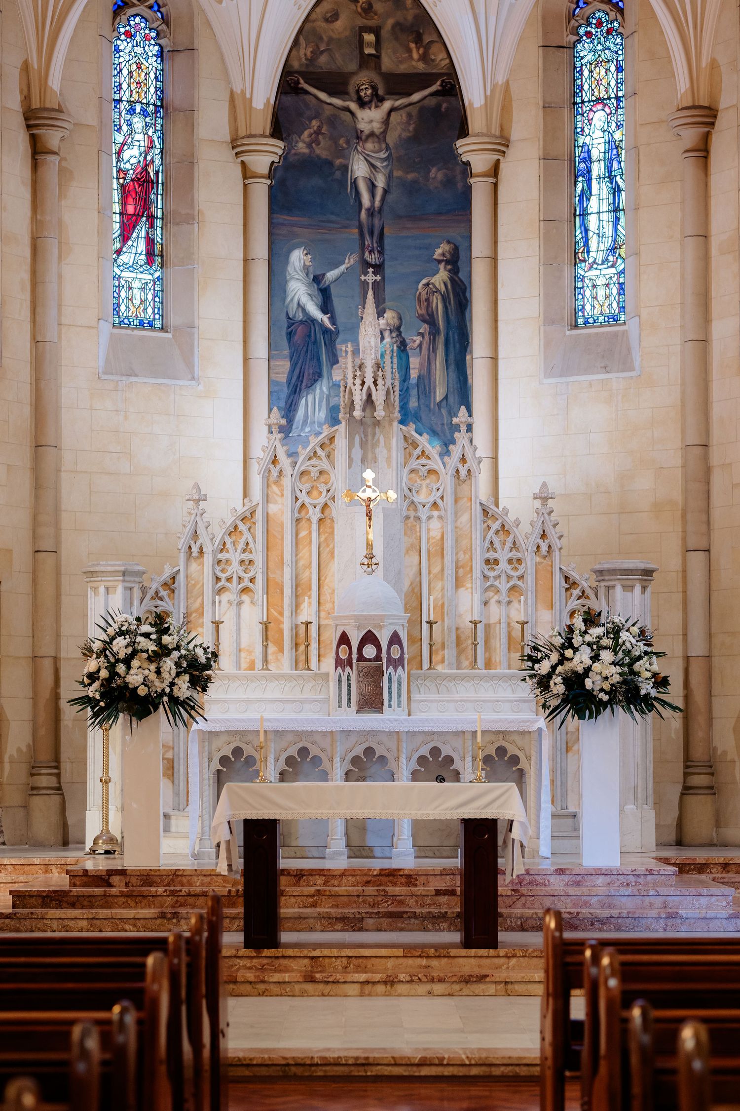 Gothic church altar with ornate crucifix, stained glass windows, white stone arches and floral arrangements.