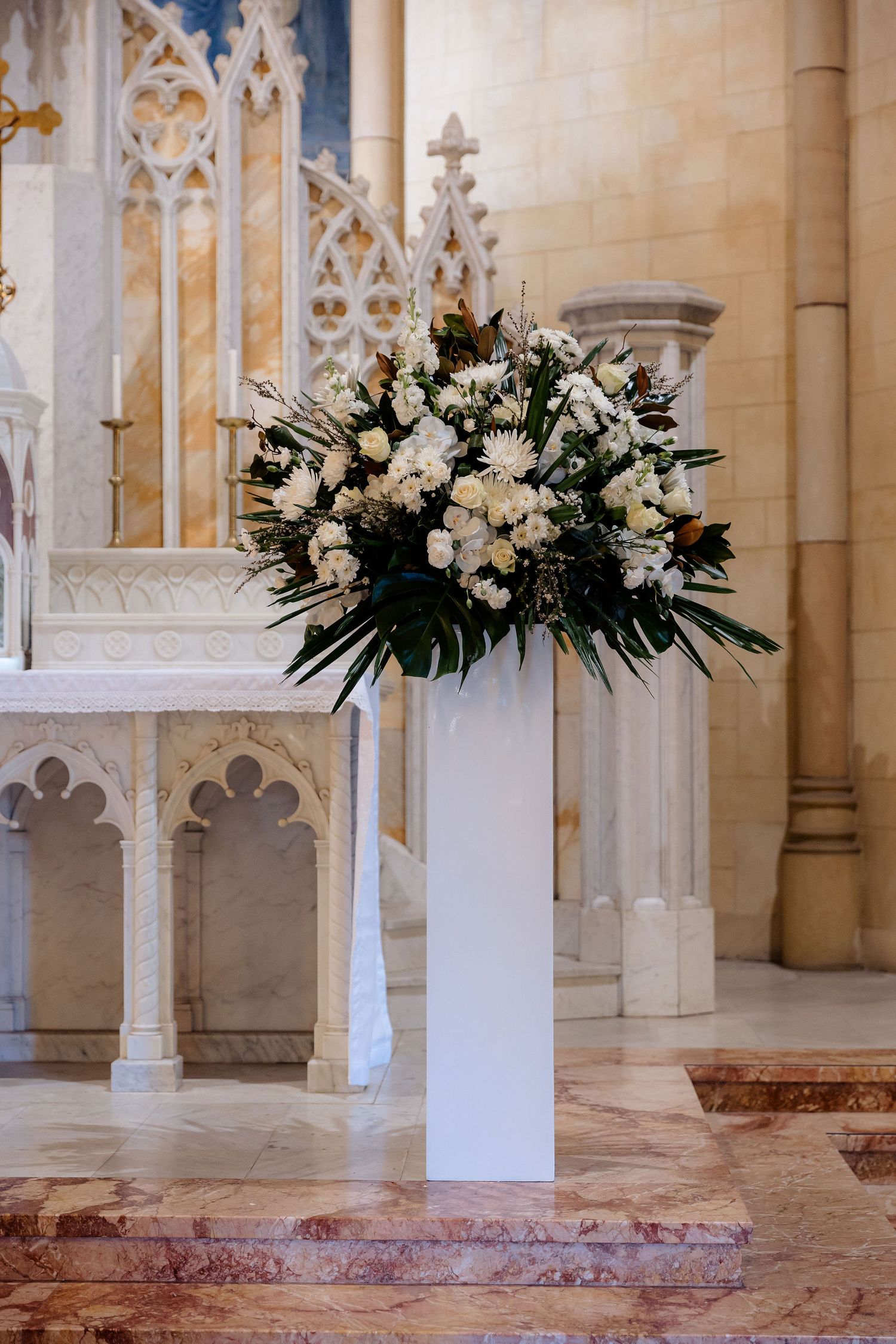 Elegant white floral arrangement on tall pedestal inside ornate Gothic church with carved stone architectural details.