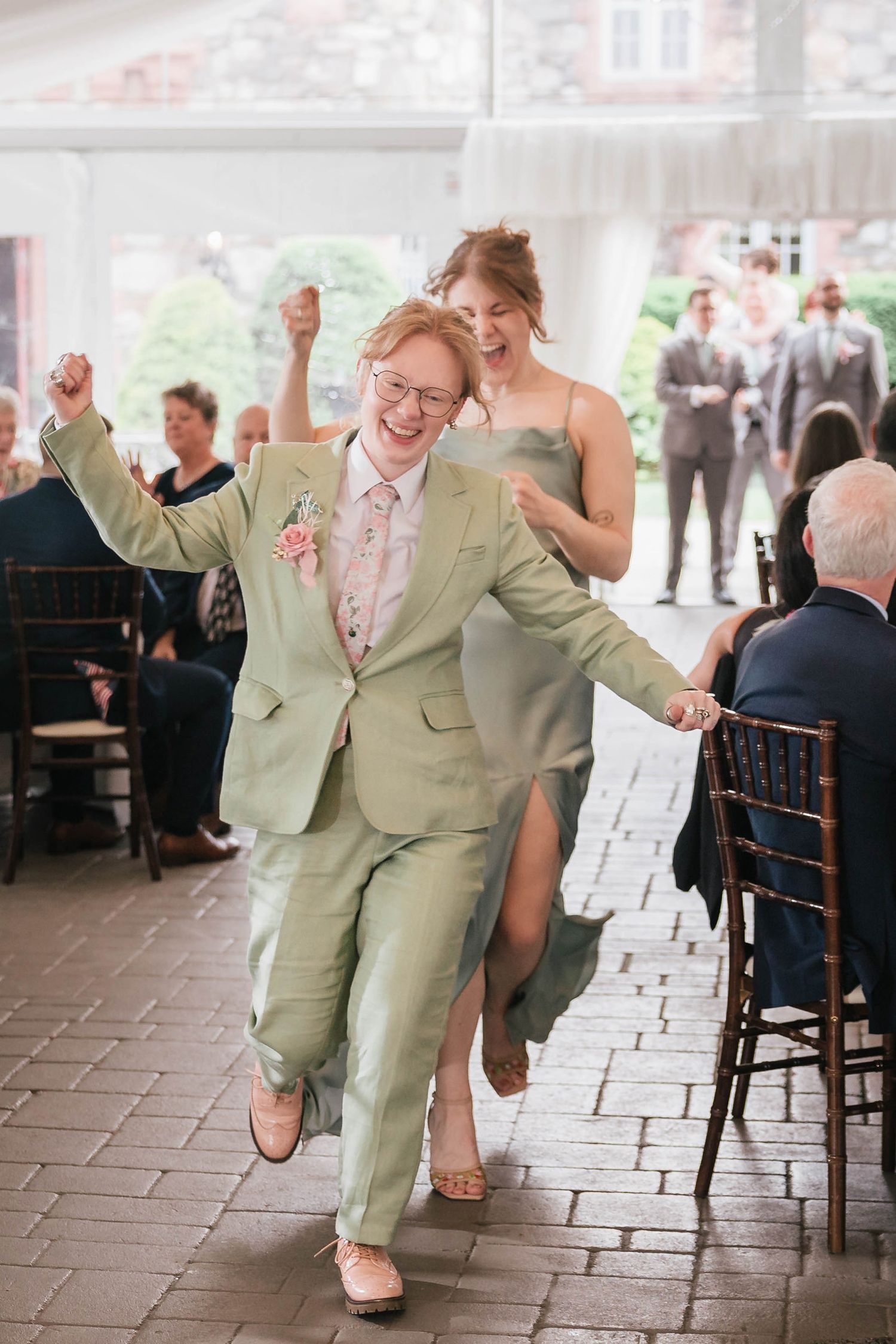 Maid of honor in a sage suit and a bridesmaid in a matching gown running into the reception tent with fists raised, cheering.