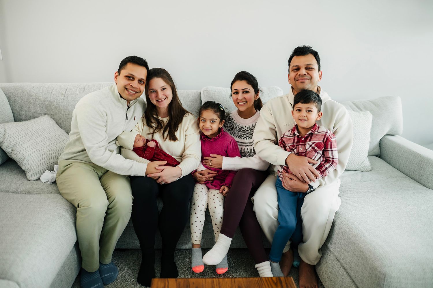 Happy family of six smiling together on a gray couch, including two young children in a bright living room.