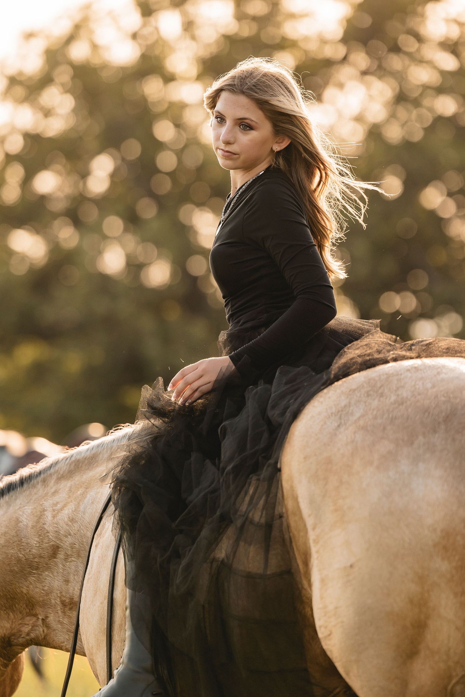 A young woman in a black dress rides a pale horse at golden hour, her hair flowing in the breeze.