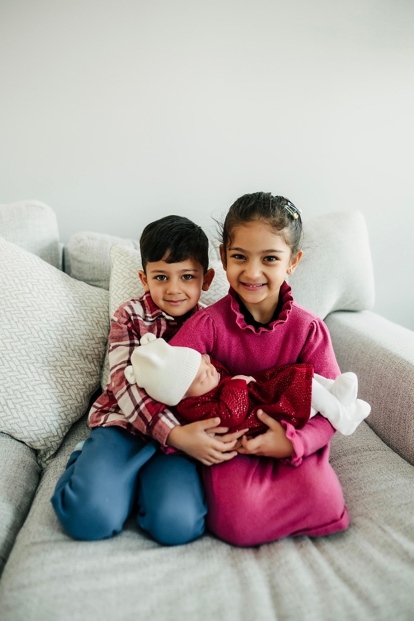 Two smiling children sit on a gray sofa holding a newborn baby wrapped in white.