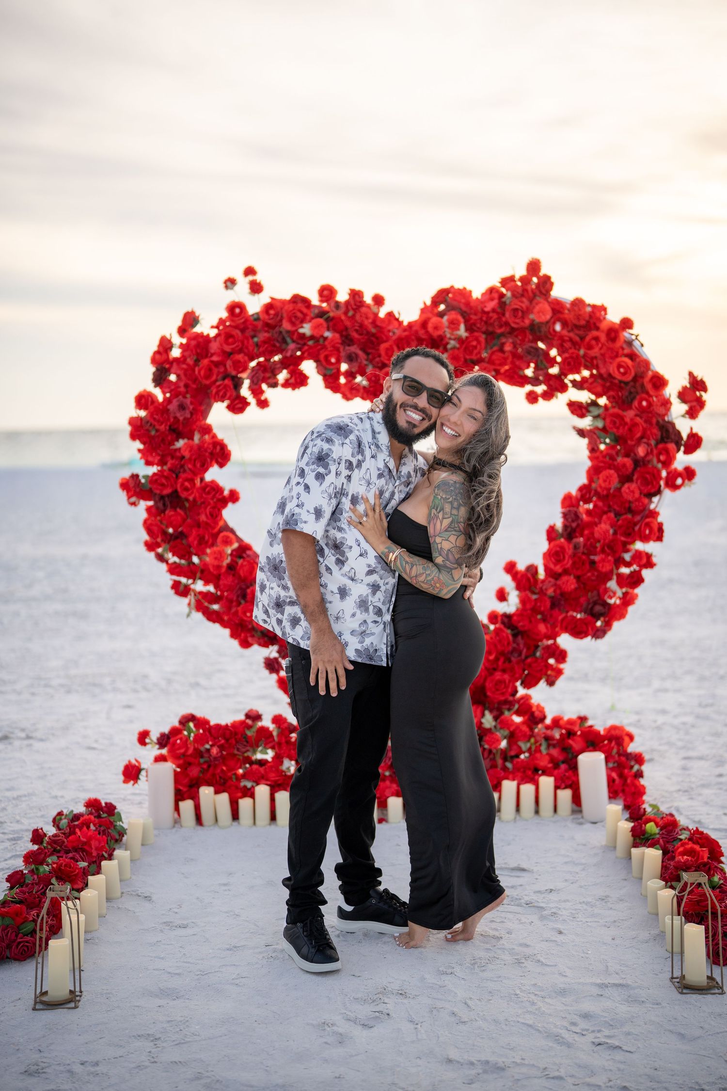 Couple embracing in front of a large red rose heart arch on a sandy beach at sunset with candles.