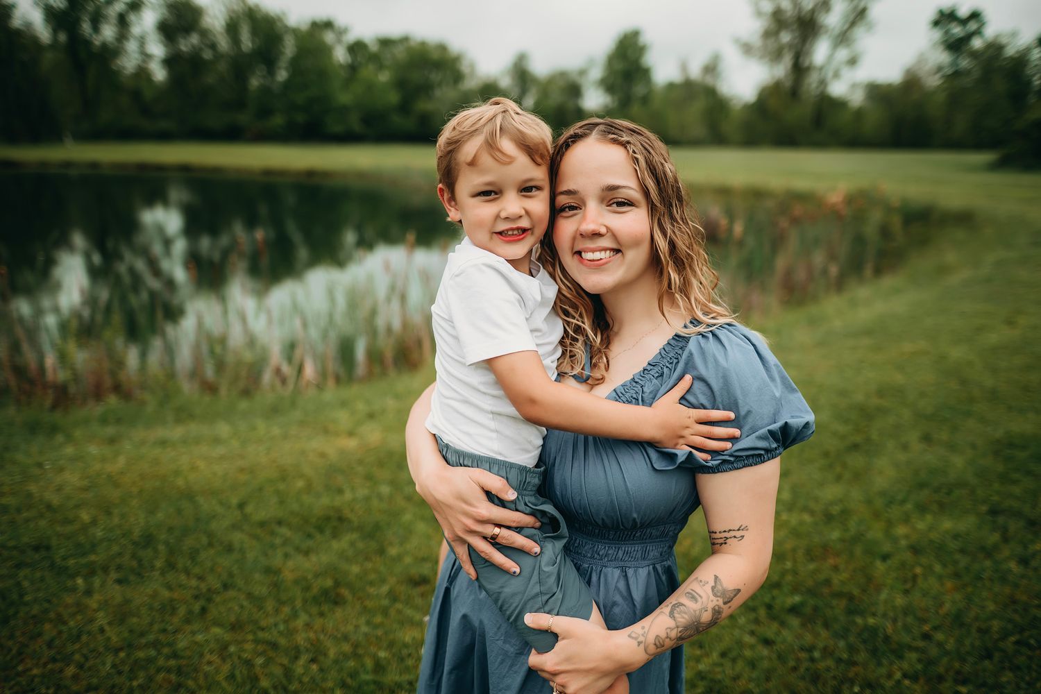 A smiling woman in a blue dress holds a young boy outdoors near a pond surrounded by green grass and trees.