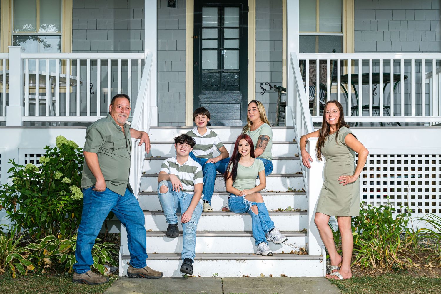 Family of six posing on white porch steps of a gray house, smiling outdoors in casual attire.