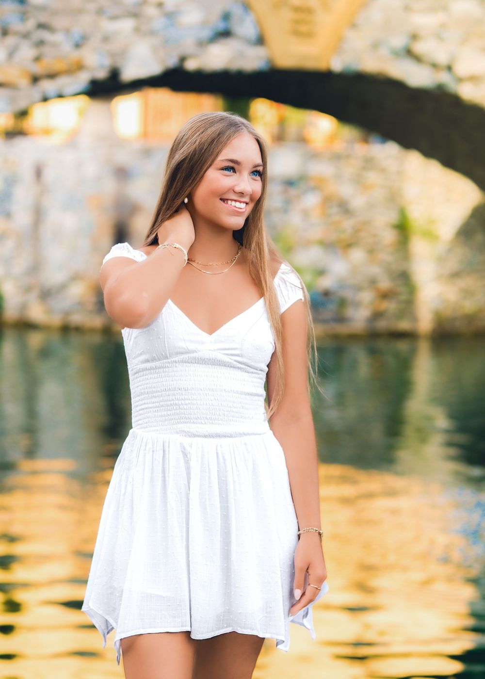 Young woman in white dress smiling near golden reflective water with stone arch bridge backdrop.