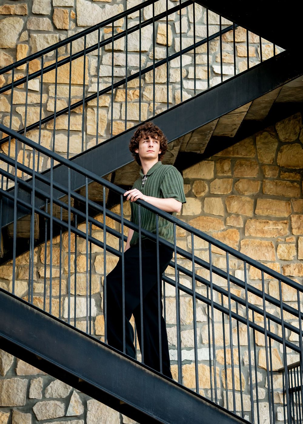 Young man with curly hair stands on metal staircase against stone wall, wearing green shirt and black pants.