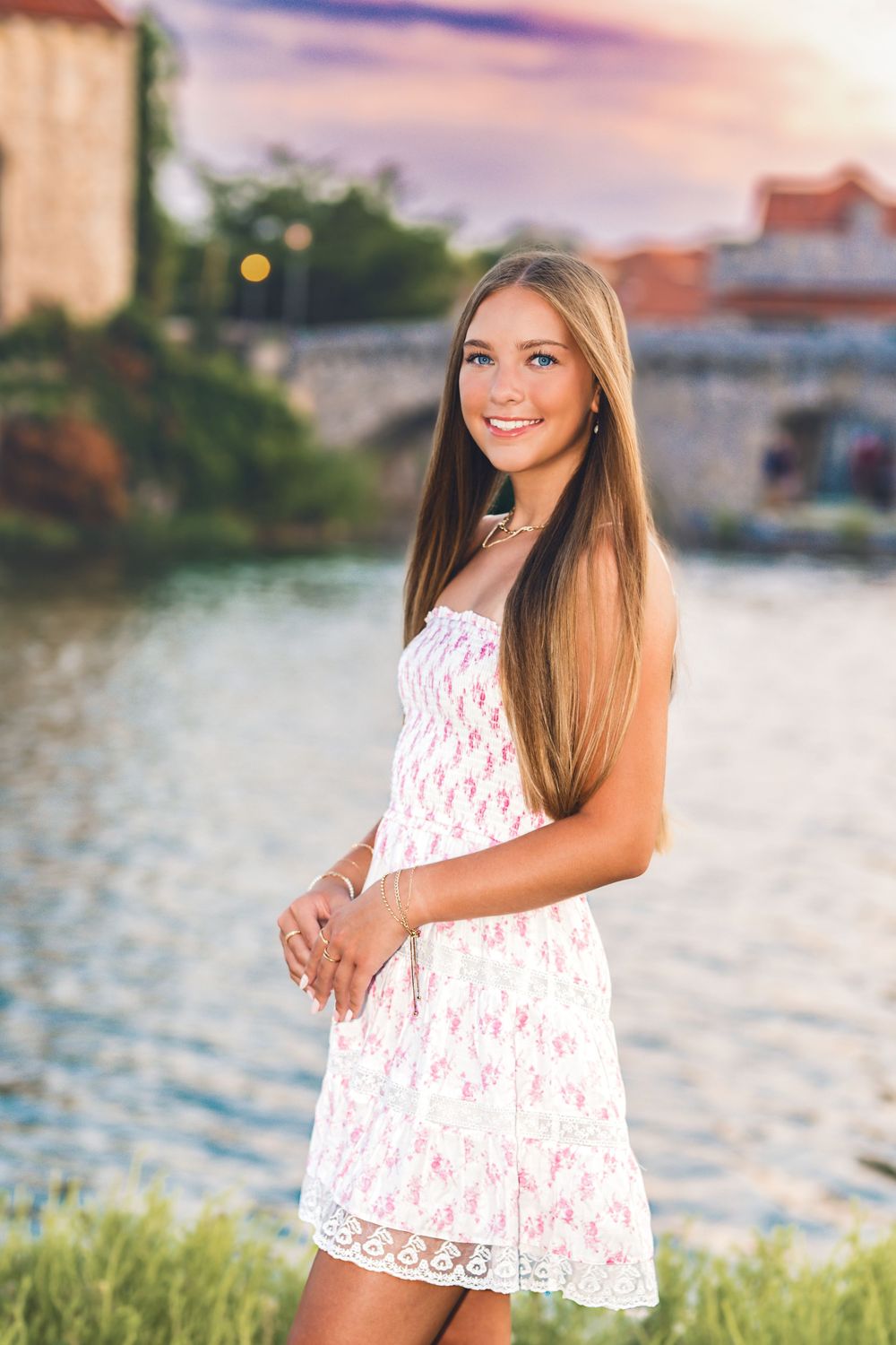Young woman in white floral sundress smiling by a river at sunset with historic buildings in background.