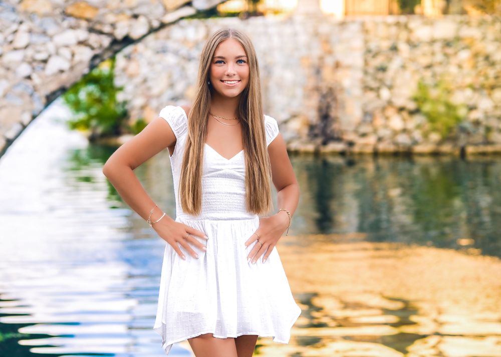 Young woman in white dress posing confidently by a reflective waterway with a stone arch bridge backdrop.