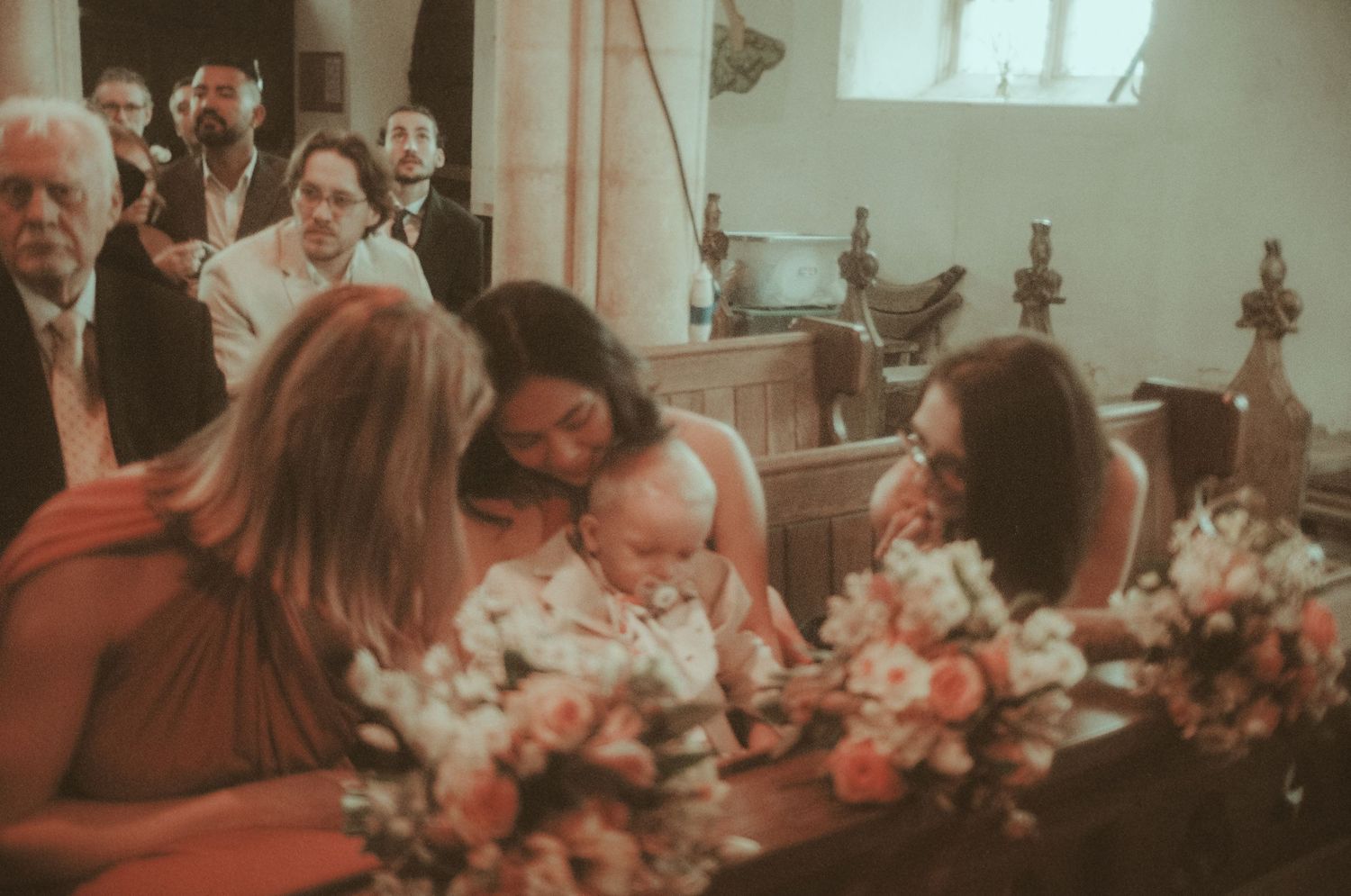 Women in coral dresses admire a baby at a church wedding ceremony, surrounded by floral arrangements.