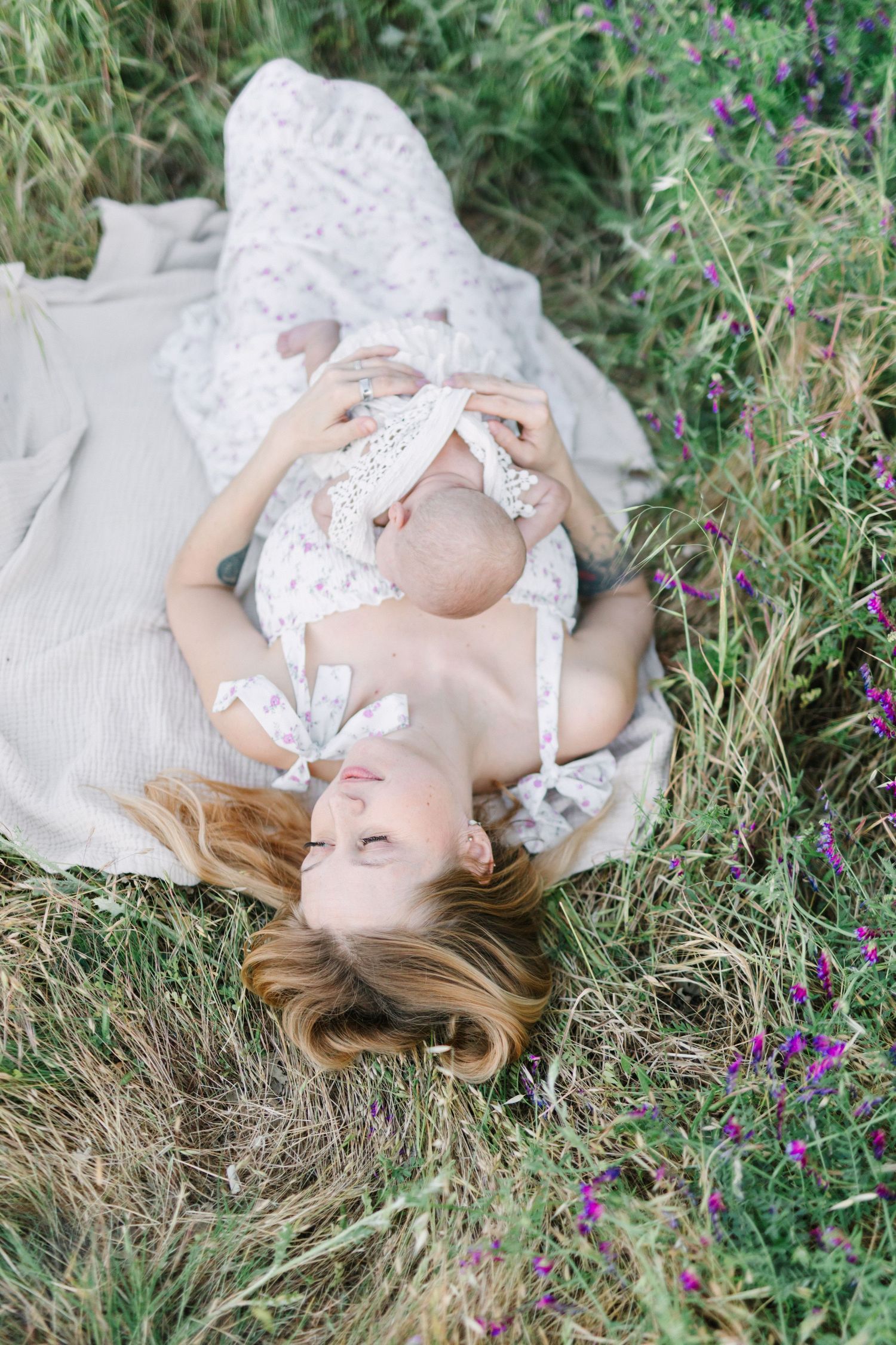 A mother lies in a wildflower meadow on a blanket, cradling her newborn baby while wearing a white lace dress.