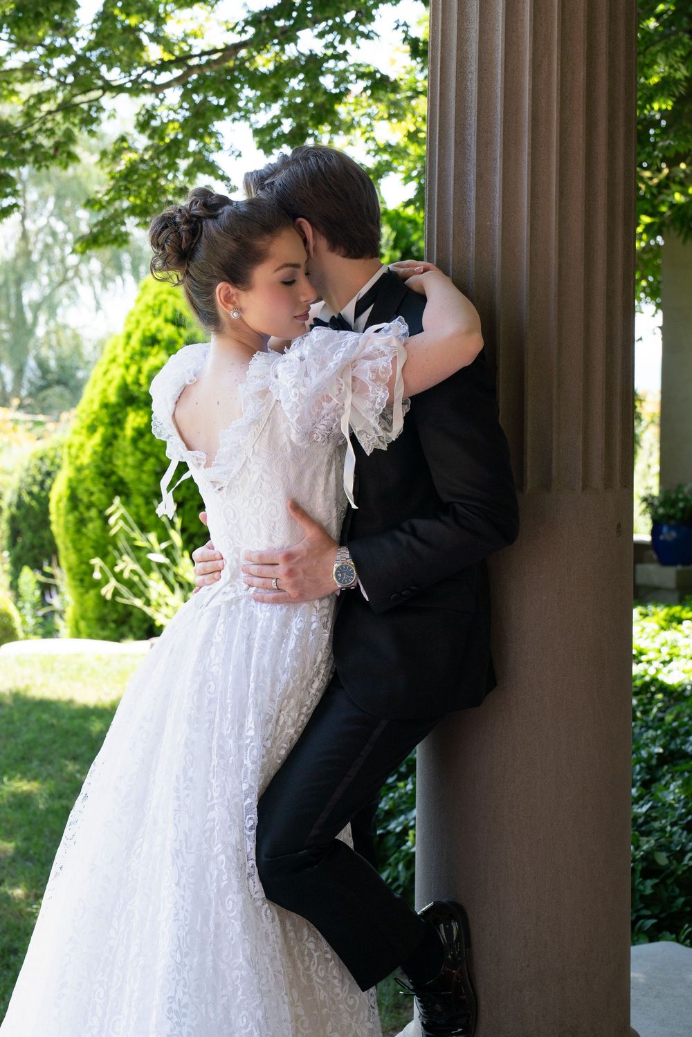 Bride and groom share an intimate embrace against a column in a lush garden, she in lace gown, he in black tuxedo.