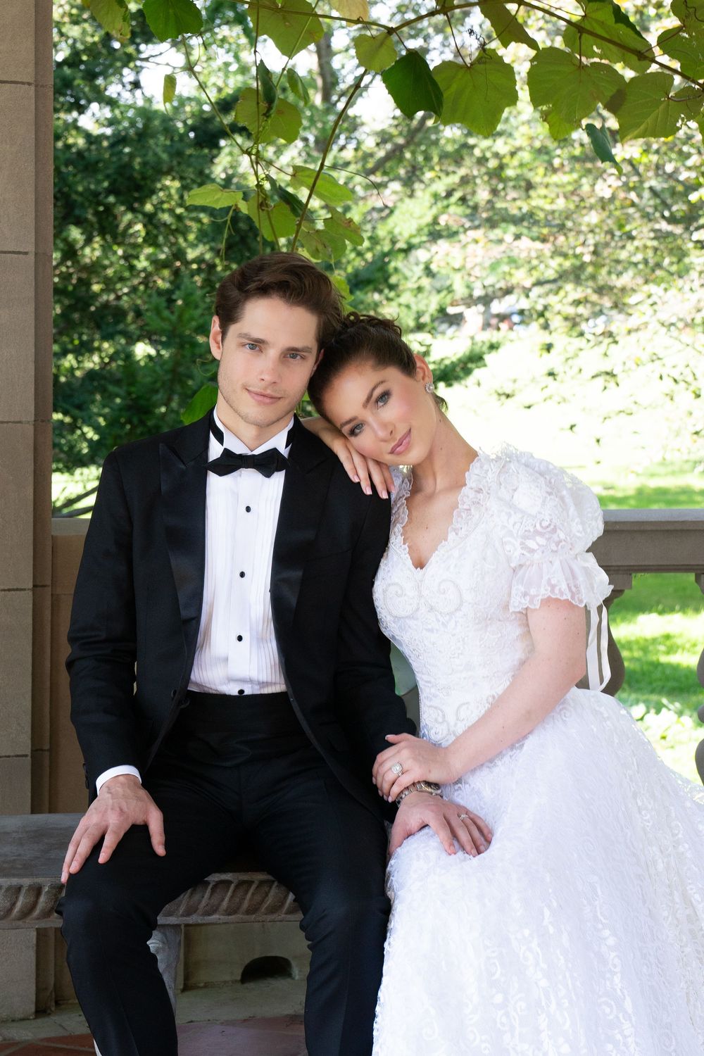 Wedding couple sits together on stone steps under green foliage, bride in white lace ballgown, groom in black tuxedo.