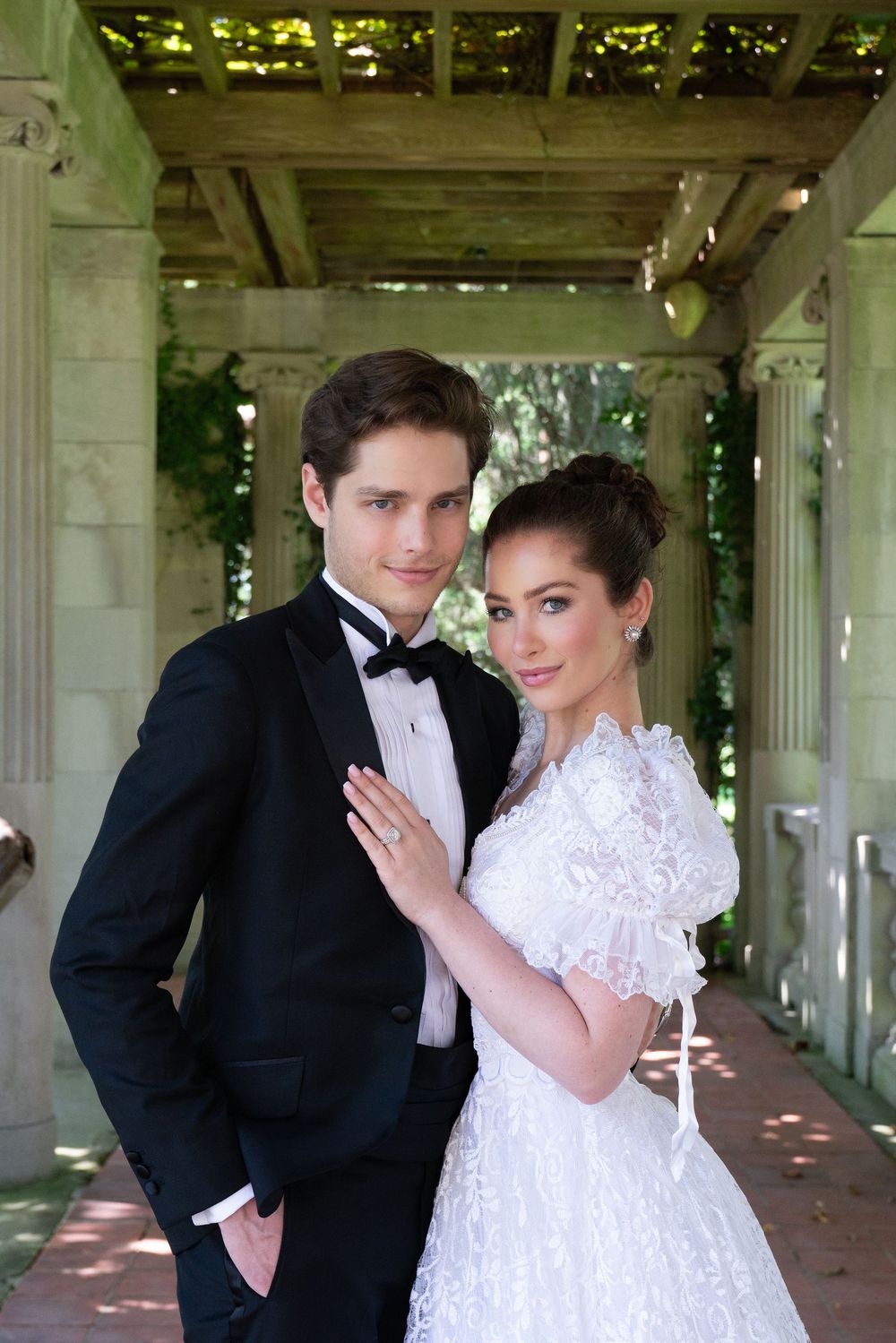 Elegant wedding couple poses under a stone pergola, bride in lace gown with puff sleeves, groom in classic black tuxedo.