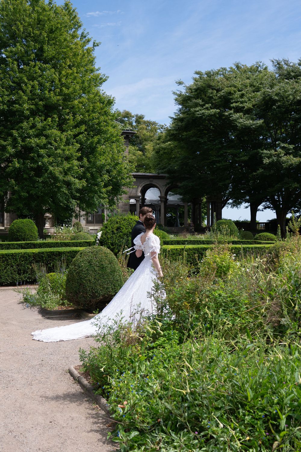 Bride and groom walking through formal garden with long white train trailing on path.