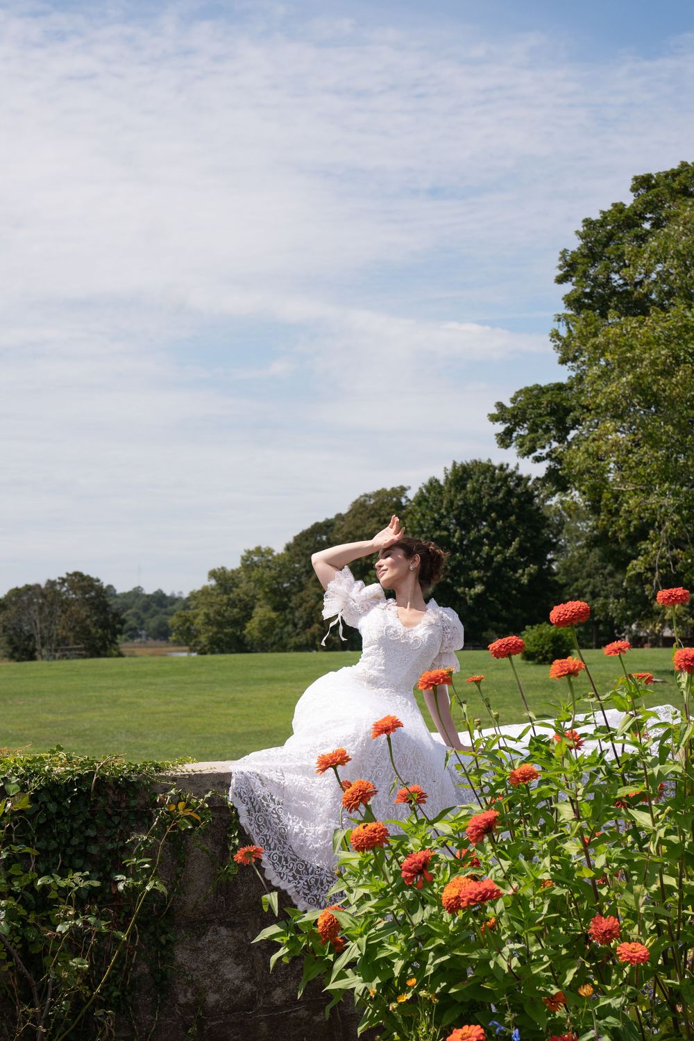 Bride in white lace gown sitting on stone wall surrounded by orange flowers in sunny garden.