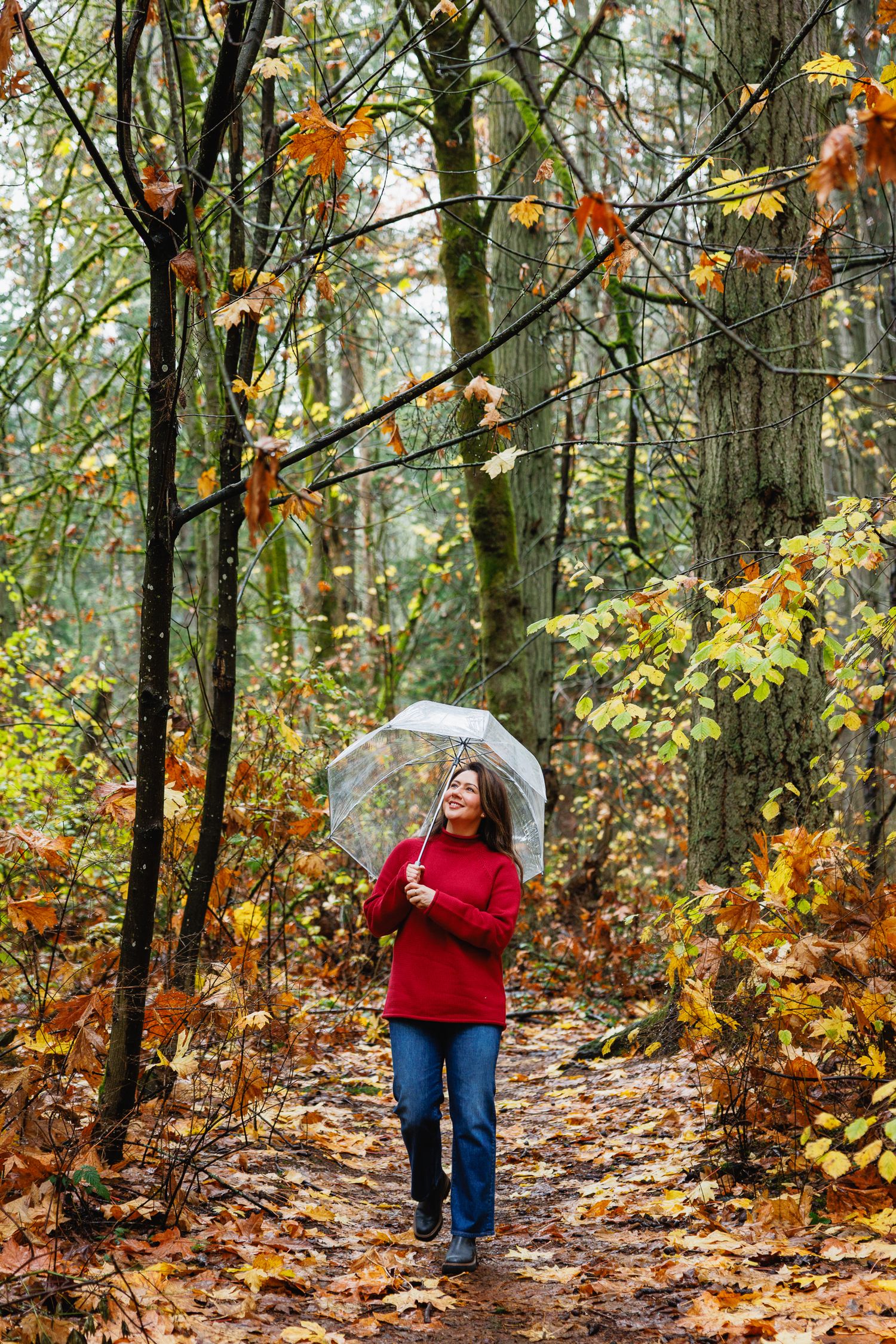 Woman in red sweater holding clear umbrella walks on leaf-covered forest path in autumn.