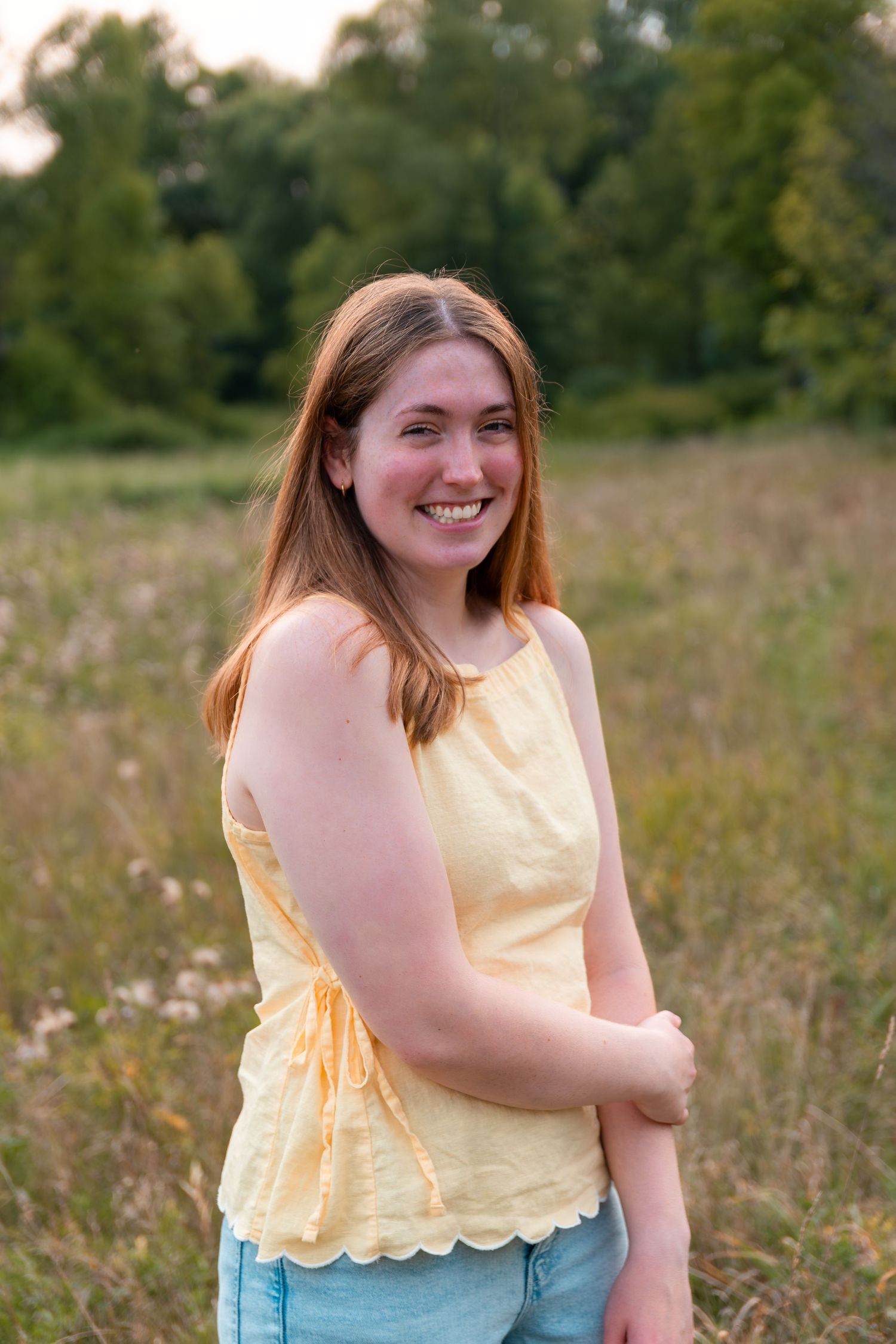 Smiling young woman with brown hair wearing a yellow sleeveless top and blue shorts standing in a green meadow.