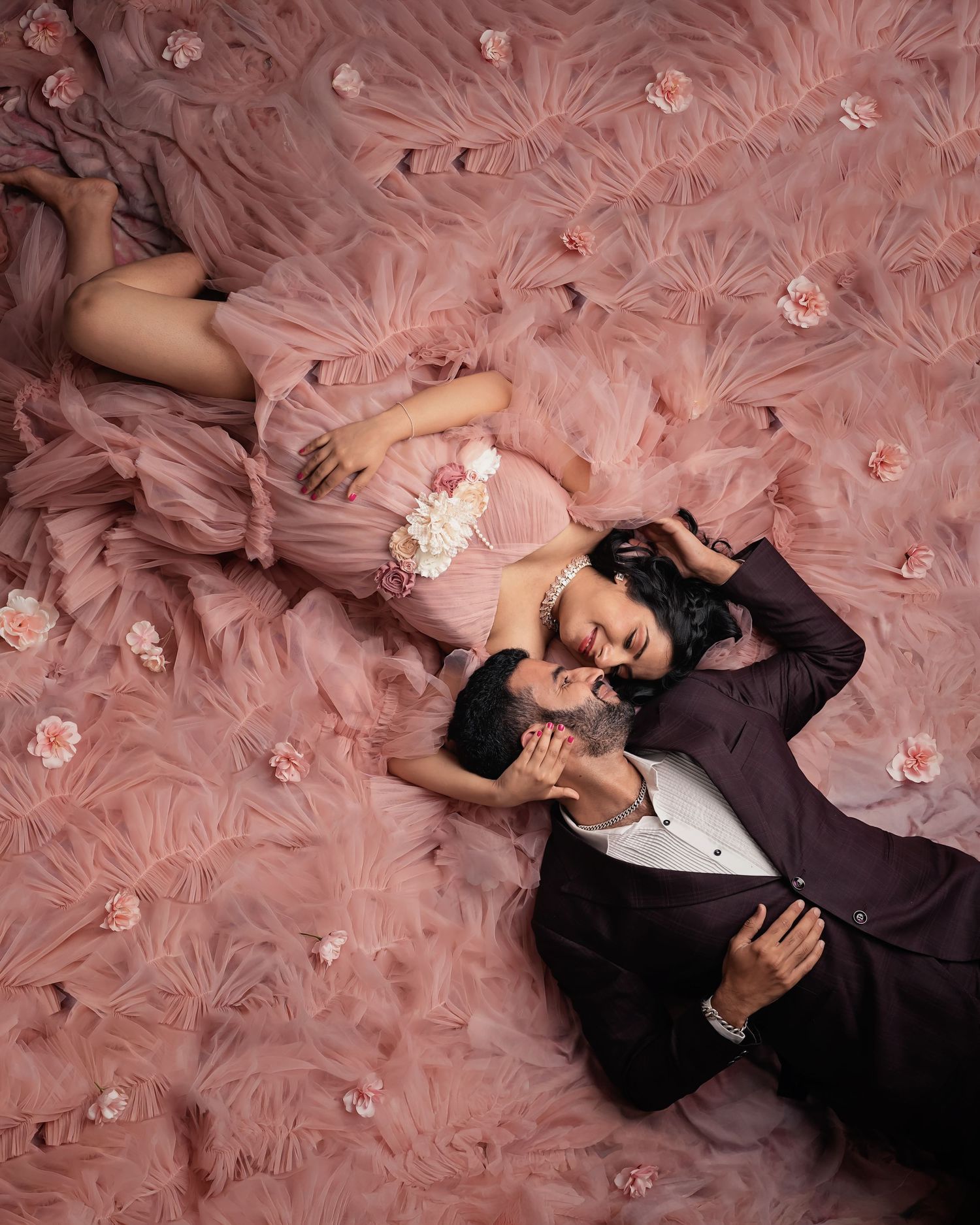 Couple in elegant attire lying on a pink ruffled gown with floral accents in a romantic styled photo shoot.