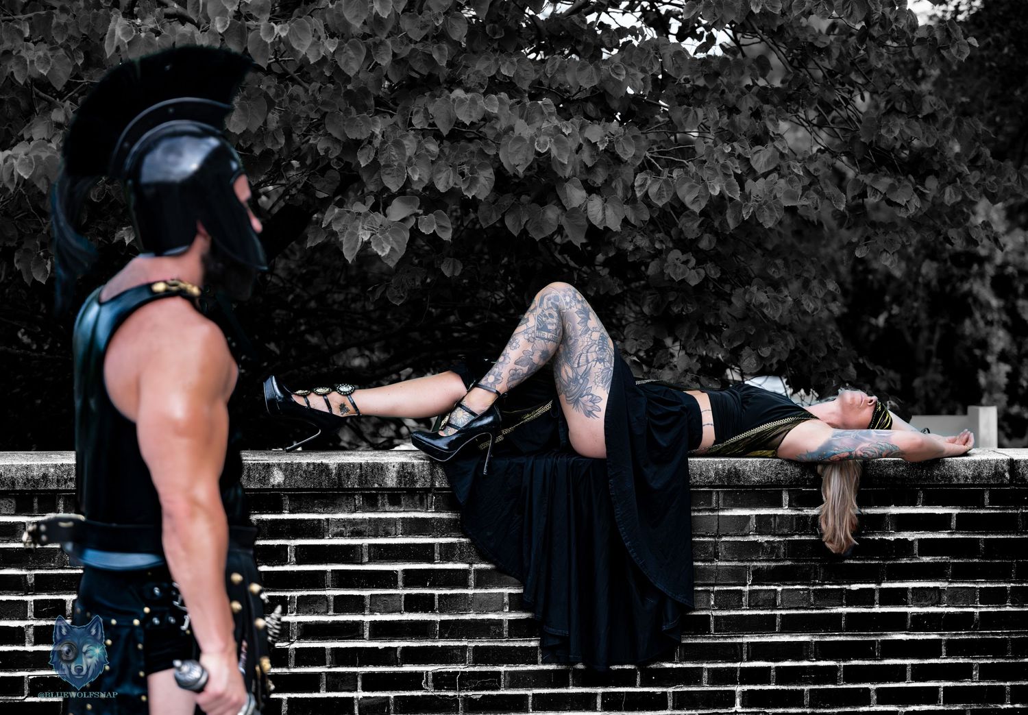 Spartan cosplayer watches tattooed woman in black dress reclining on brick wall outdoors. Chi.Shooters meet tonybrettman tattoo_jessie