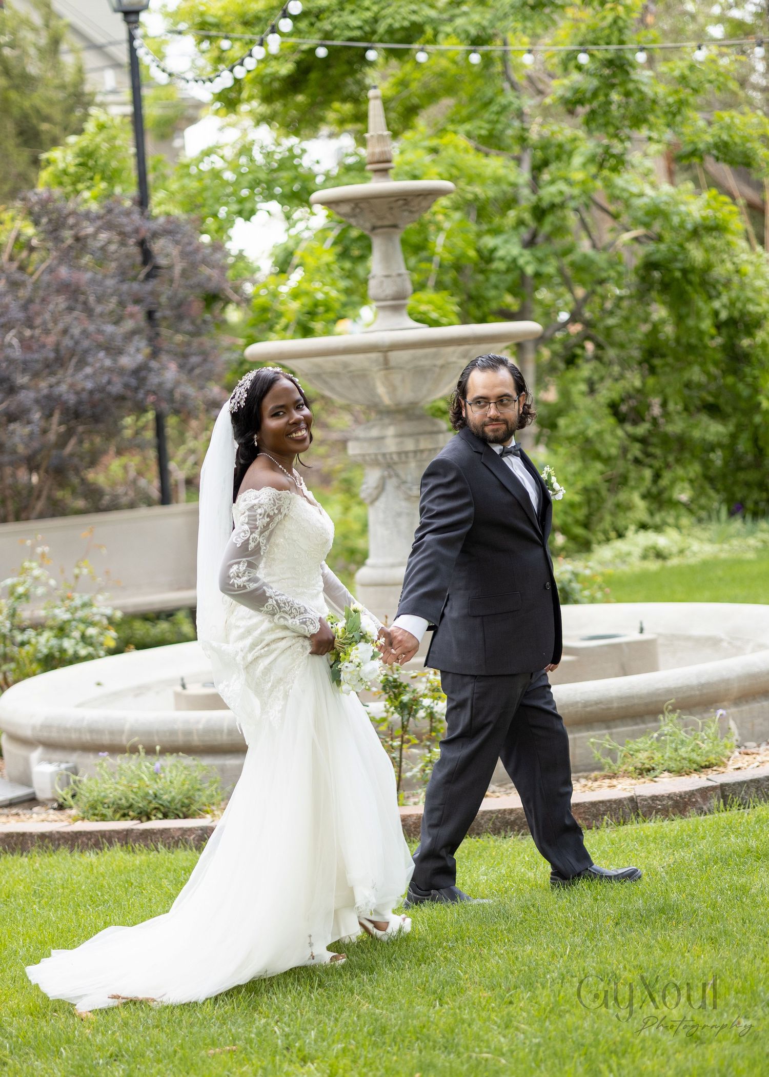 Bride in white lace gown and groom in black tuxedo hold hands by stone fountain in lush garden wedding photo.