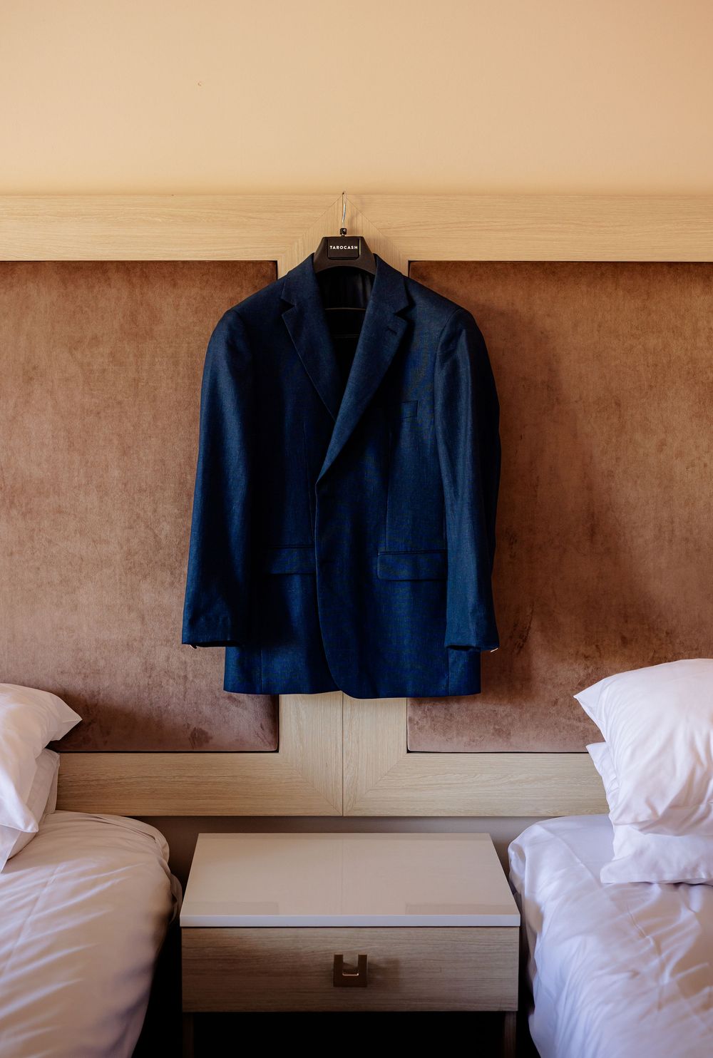 Navy blue suit jacket hanging on a hanger between two single beds with white pillows in a hotel room.