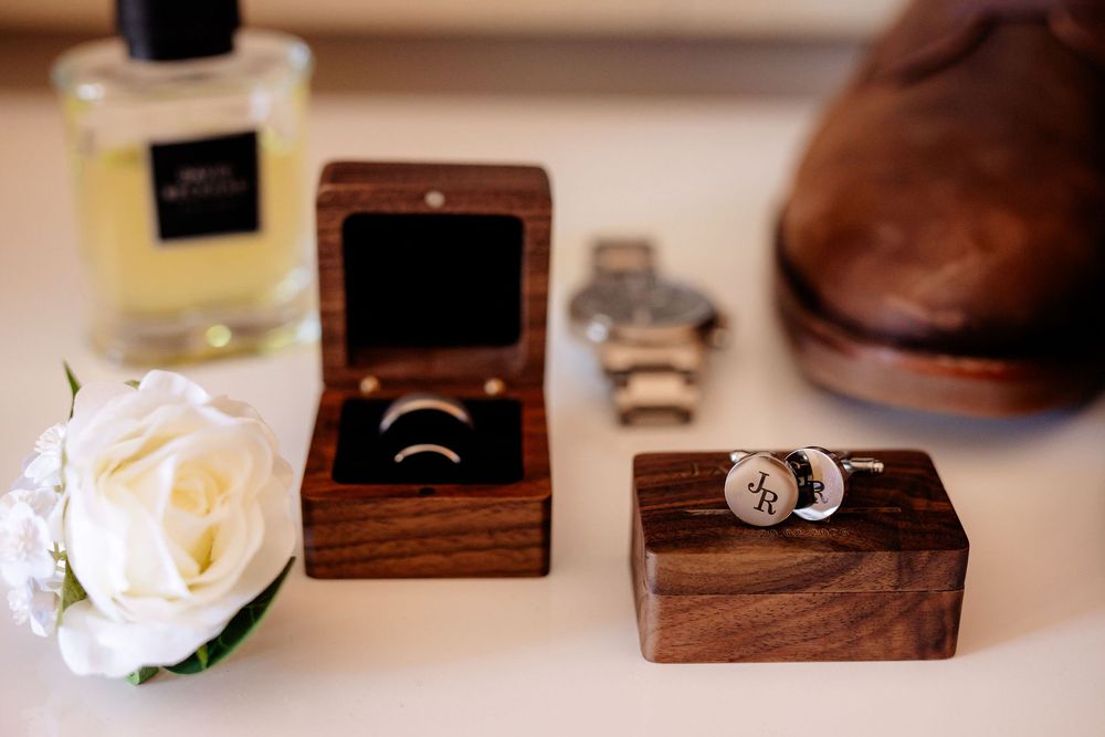 Elegant wedding flat lay featuring gold rings in wooden box, cufflinks, watch, white rose, cologne, and brown leather shoe.