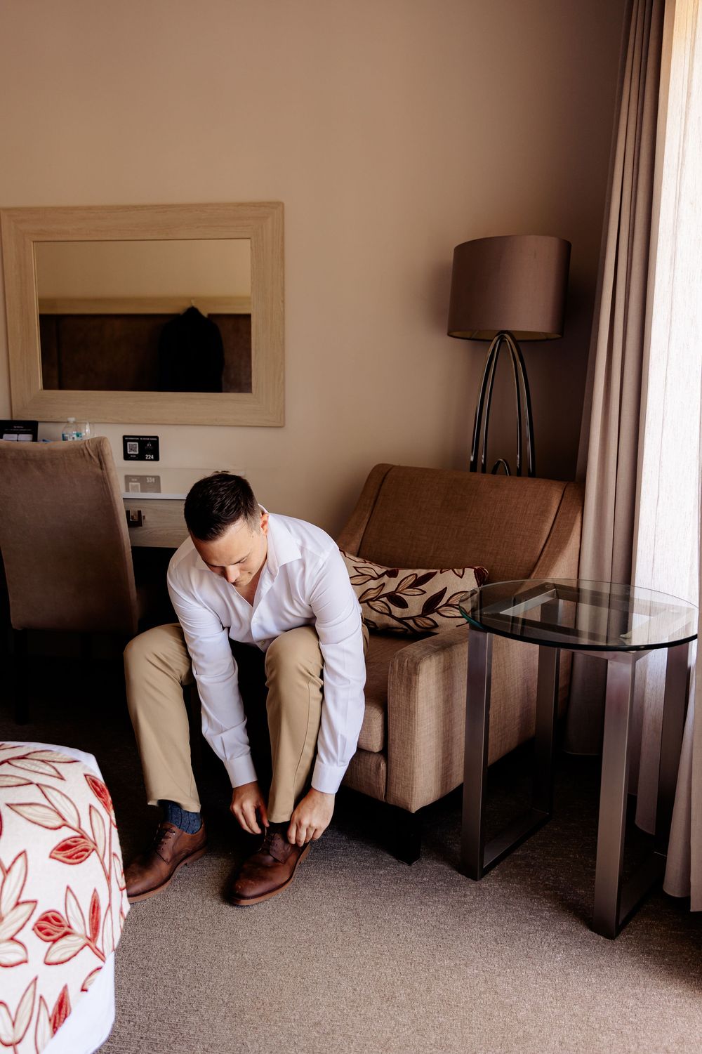 A man in a white shirt and khaki pants sits in a hotel room armchair, tying his brown dress shoes.