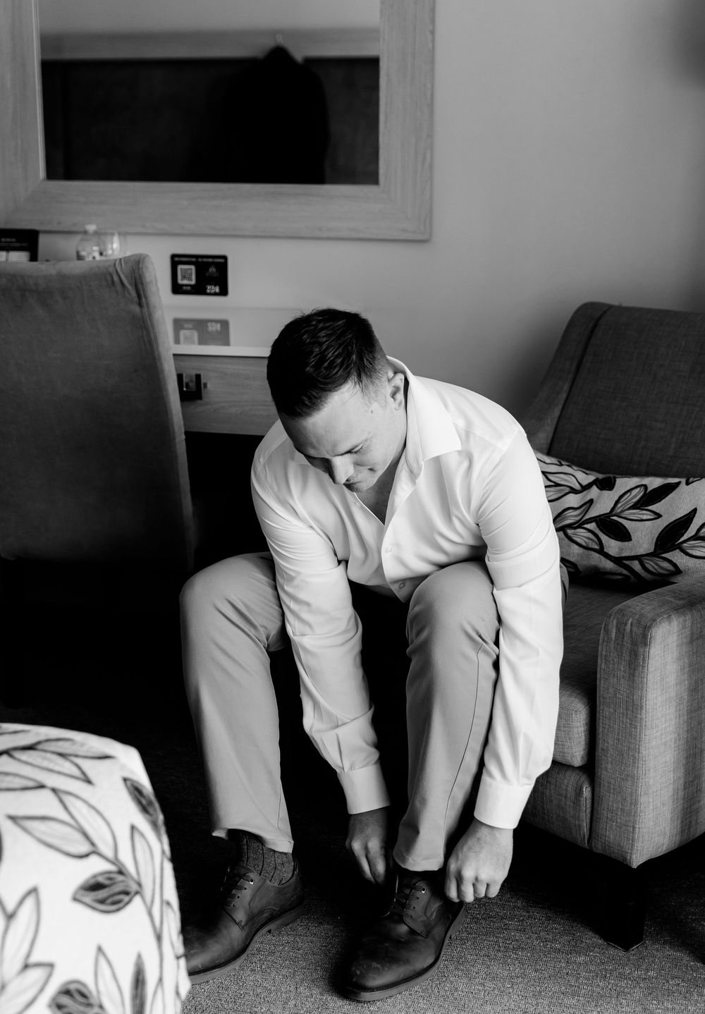 Black and white photo of a groom in a white shirt sitting on a sofa putting on his dress shoes before his wedding.
