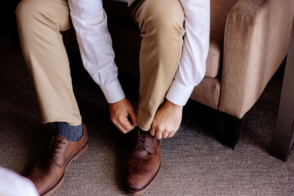 Man in khaki pants tying brown leather dress shoes while seated, wearing white shirt and dark socks.