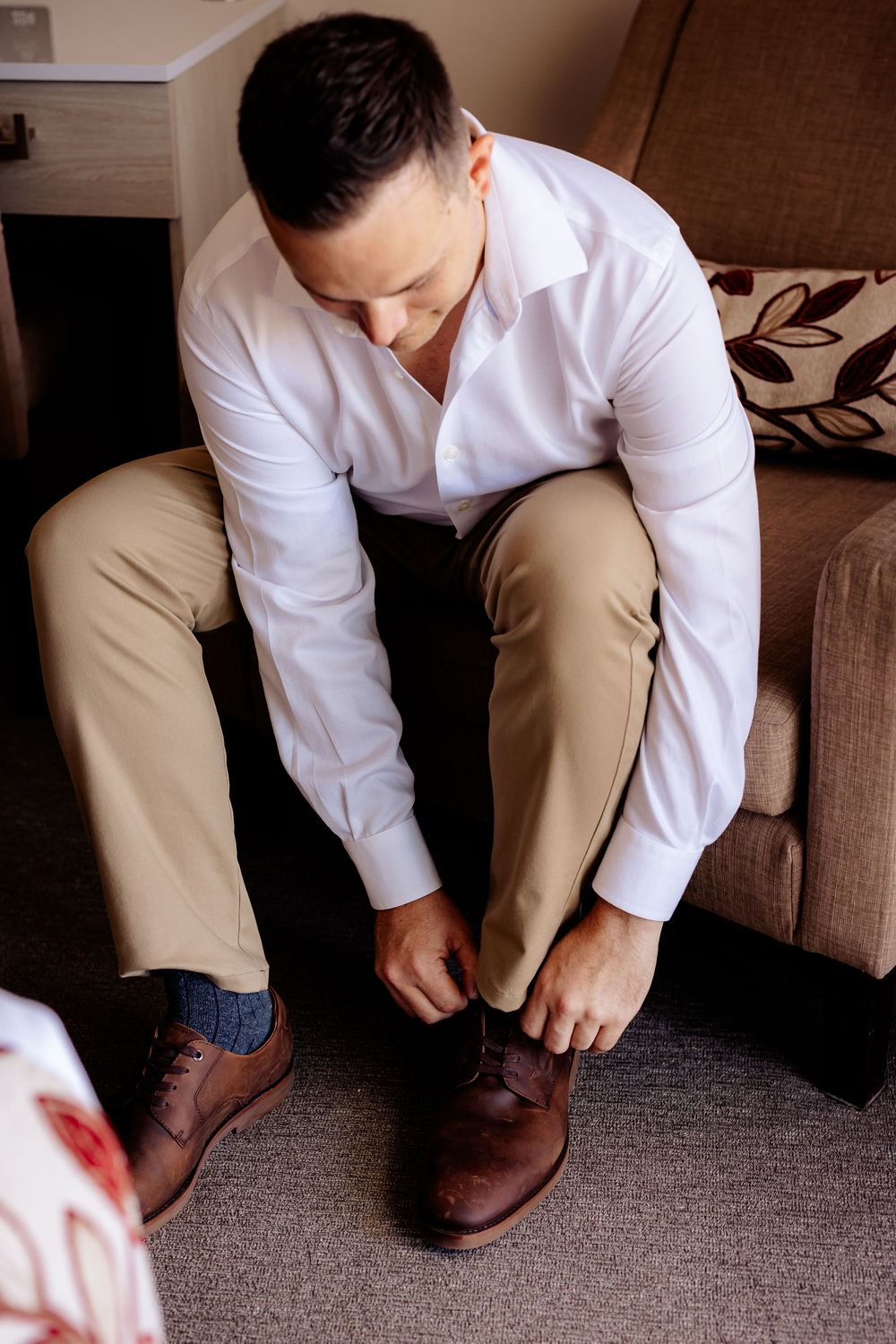 Man in white shirt and khaki pants tying brown dress shoes while seated on a sofa.