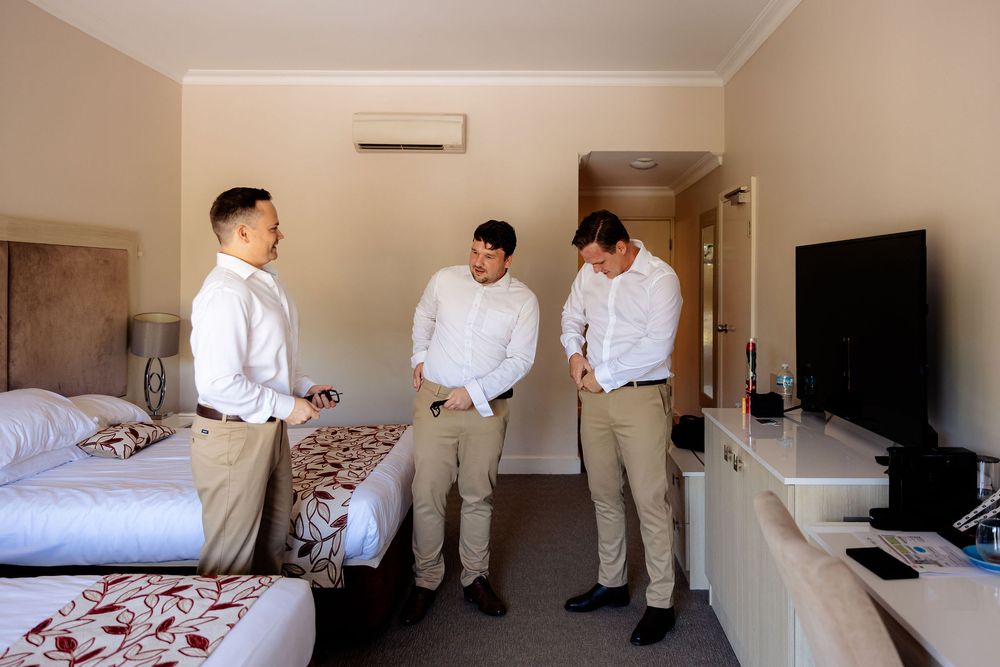 Three groomsmen in white shirts and khaki pants getting ready in a hotel room before a wedding ceremony.