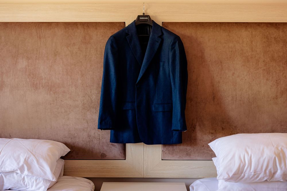 Navy blue suit jacket hanging on a hanger between two white pillows against a textured beige wall.