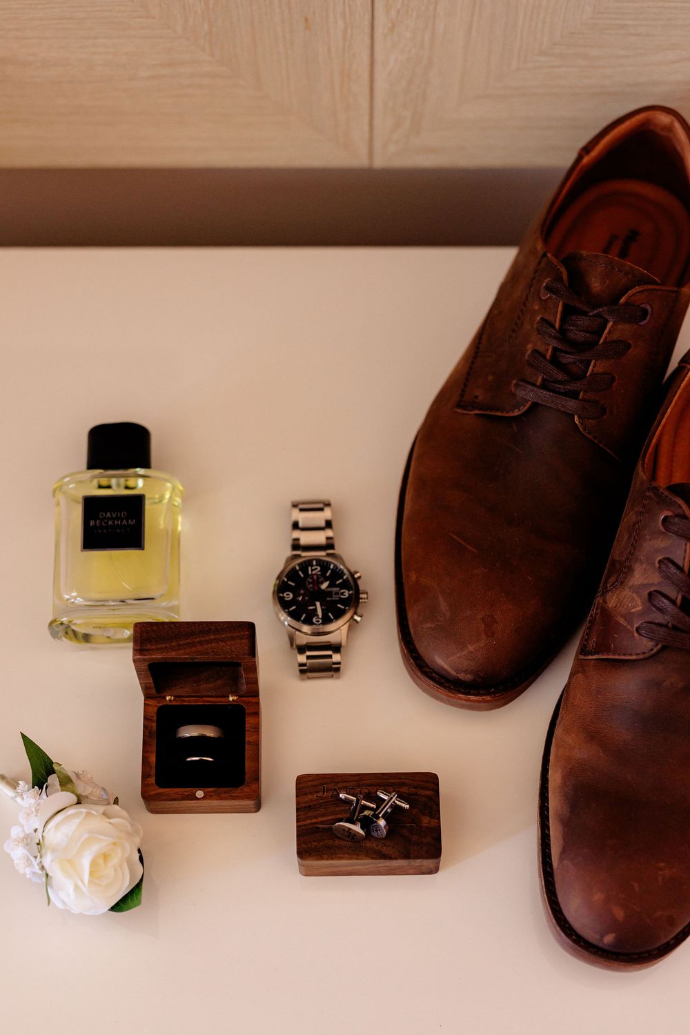 Groom wedding flat lay with brown leather dress shoes, silver watch, cologne, ring box, cufflinks, and white flower.
