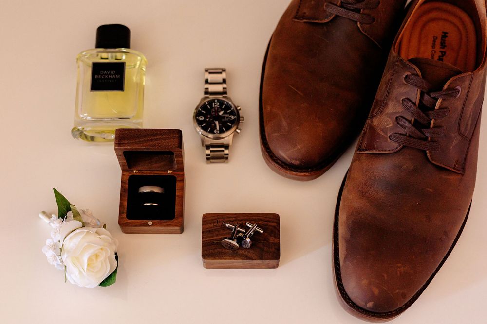 Brown leather dress shoes with a watch, cologne, wedding rings in a wooden box, cufflinks, and a white boutonniere on a neutral background.