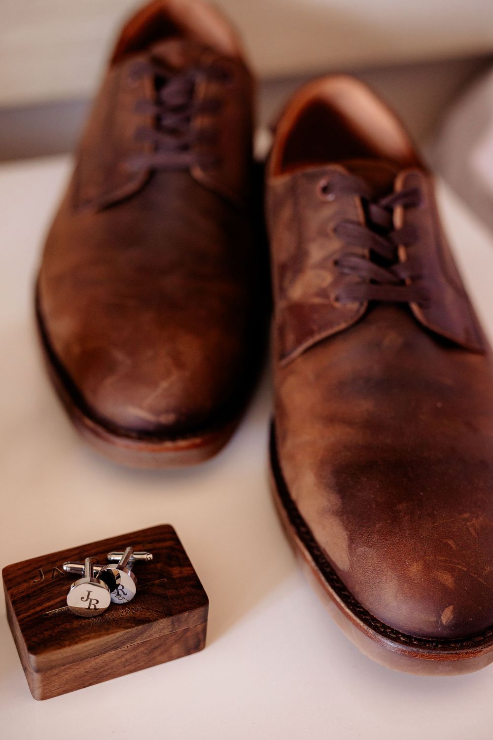 Brown leather oxford dress shoes beside silver monogrammed cufflinks on a wooden box, groom wedding accessories.