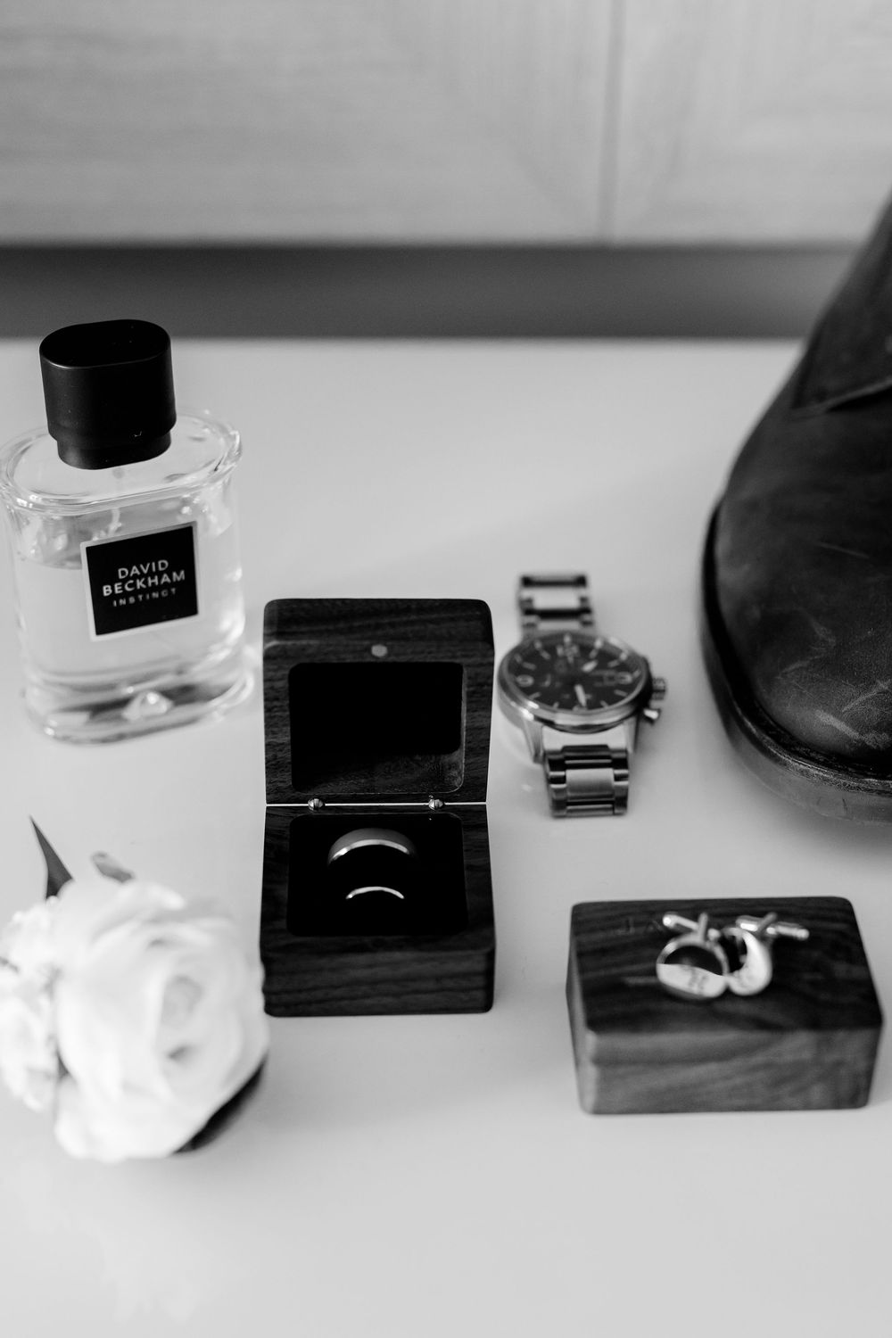 Black and white flat lay of groom wedding accessories including David Beckham cologne, ring box, watch, cufflinks, and dress shoe.