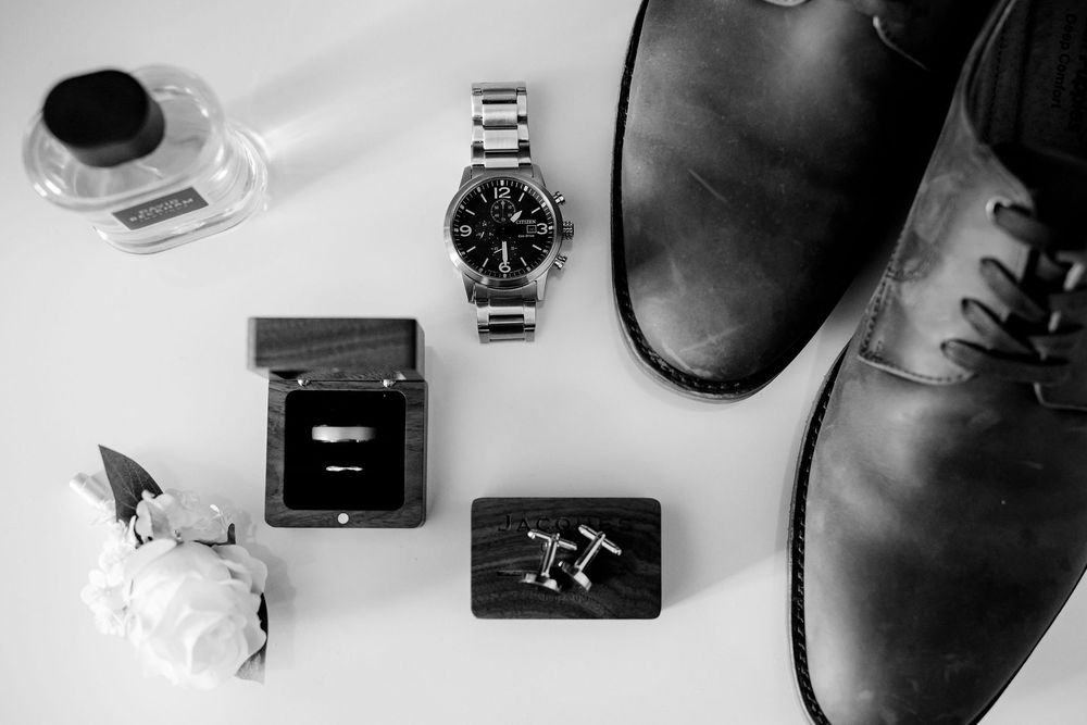 Black and white flat lay of groom accessories including watch, cufflinks, ring box, leather shoes, and cologne.