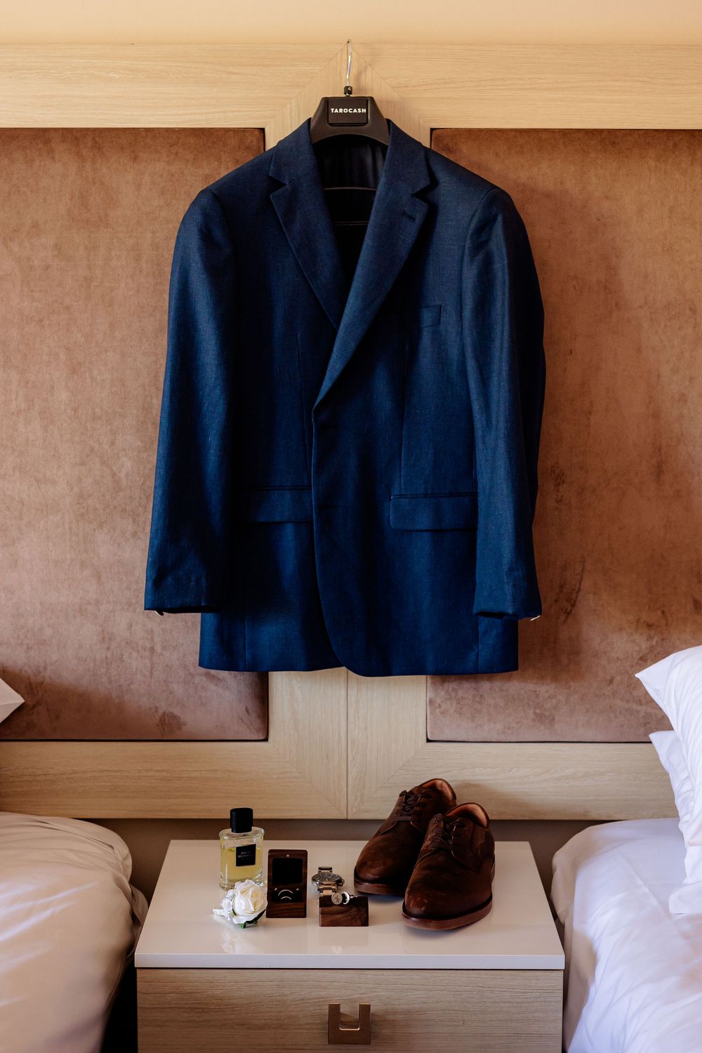 Navy blue suit jacket hanging on hanger above nightstand with brown dress shoes, cologne, and accessories in hotel room.
