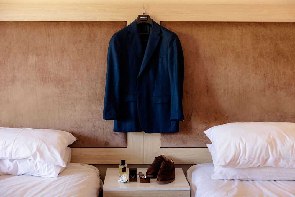 Navy blue suit jacket hanging between two beds with white pillows and brown shoes on nightstand.
