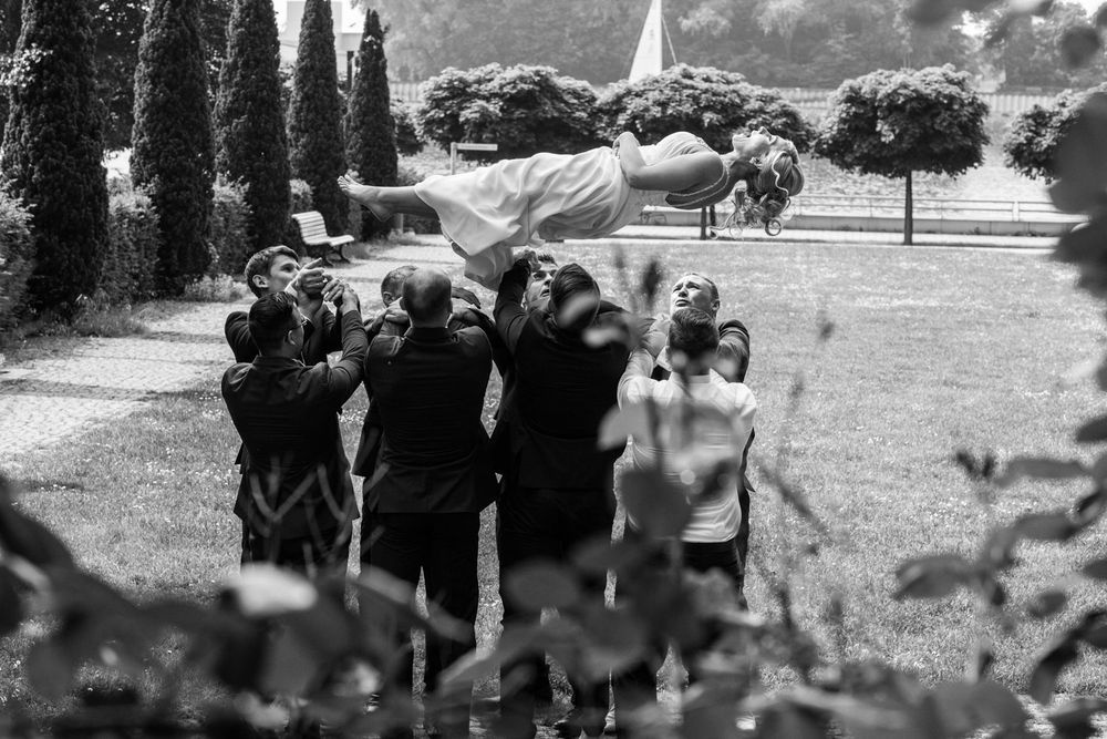 Groomsmen tossing bride in the air during outdoor wedding celebration in a garden, black and white photo.